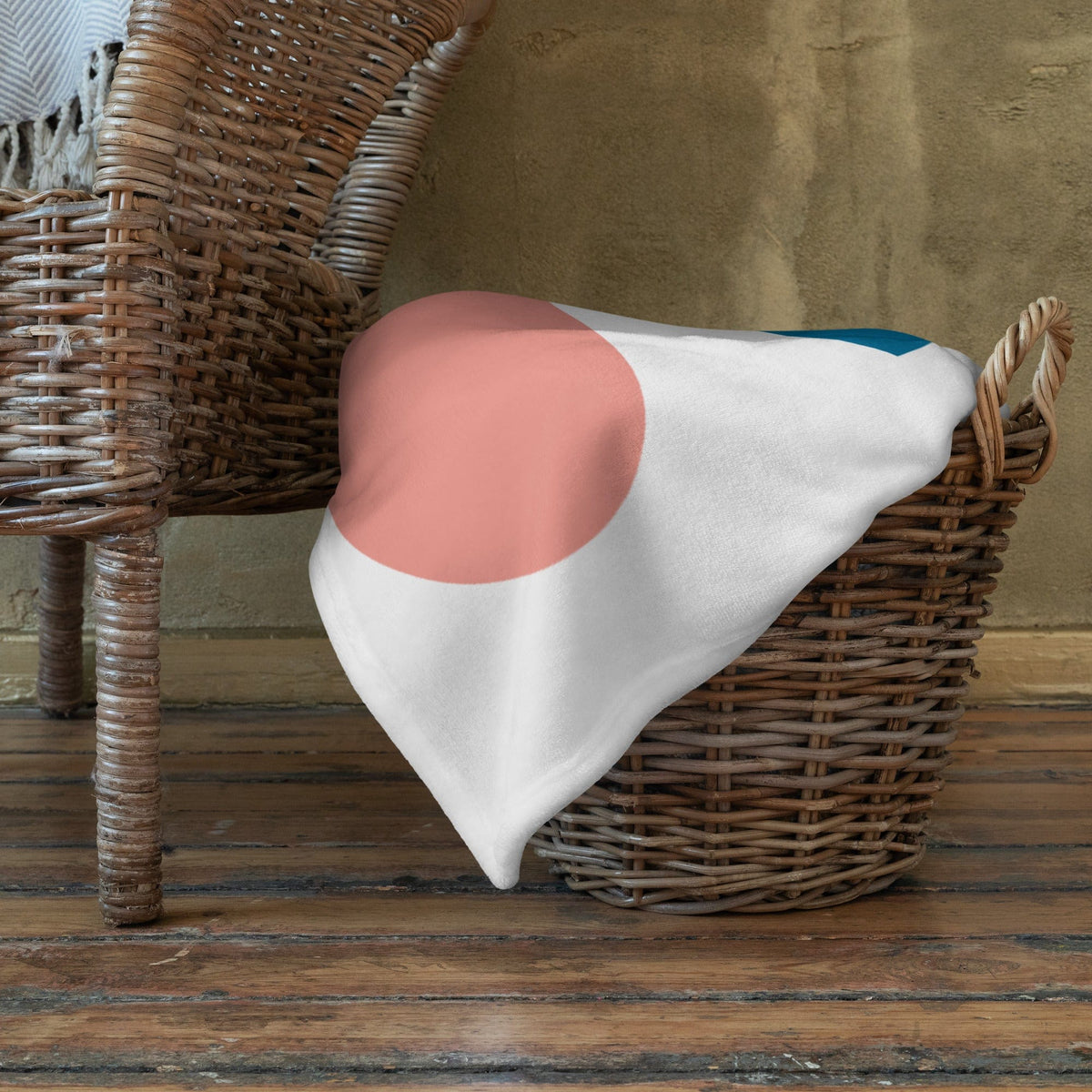 White throw blanket with large pink circle and teal accent, folded in wicker basket.